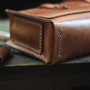 Puede incluir: Primer plano de un bolso de cuero marr&oacute;n claro hecho a mano, con costuras visibles y cierre de solapa. El bolso tiene forma rectangular y parece estar bien hecho. Un mango de madera es visible en primer plano, lo que sugiere una herramienta o accesorio.