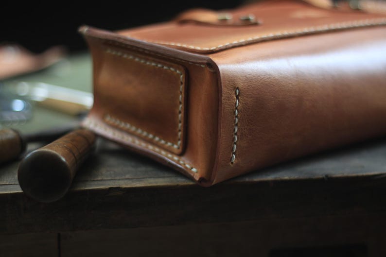 Puede incluir: Primer plano de un bolso de cuero marr&oacute;n claro hecho a mano, con costuras visibles y cierre de solapa. El bolso tiene forma rectangular y parece estar bien hecho. Un mango de madera es visible en primer plano, lo que sugiere una herramienta o accesorio.