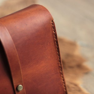 May include: Close-up of a brown leather bag with a stitched edge. The bag has a strap and a metal buckle.