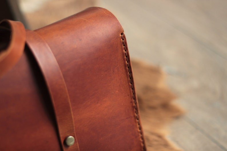 May include: Close-up of a brown leather bag with a stitched edge. The bag has a strap and a metal buckle.