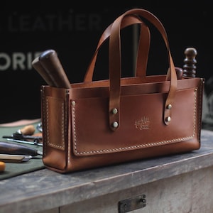 Può includere: Una borsa tote in pelle marrone con due manici e forma rettangolare. La borsa ha una tasca frontale ed è piena di attrezzi. La borsa è appoggiata su un tavolo di legno. Le parole "Canvas & Leather Accessories" sono sullo sfondo.