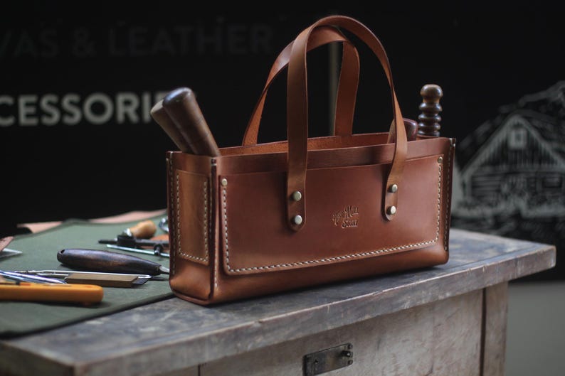 Puede incluir: Un bolso tote de cuero marr&oacute;n con dos asas y forma rectangular. El bolso tiene un bolsillo delantero y est&aacute; lleno de herramientas. El bolso est&aacute; sobre una mesa de madera. Las palabras "Canvas & Leather Accessories" est&aacute;n en el fondo.