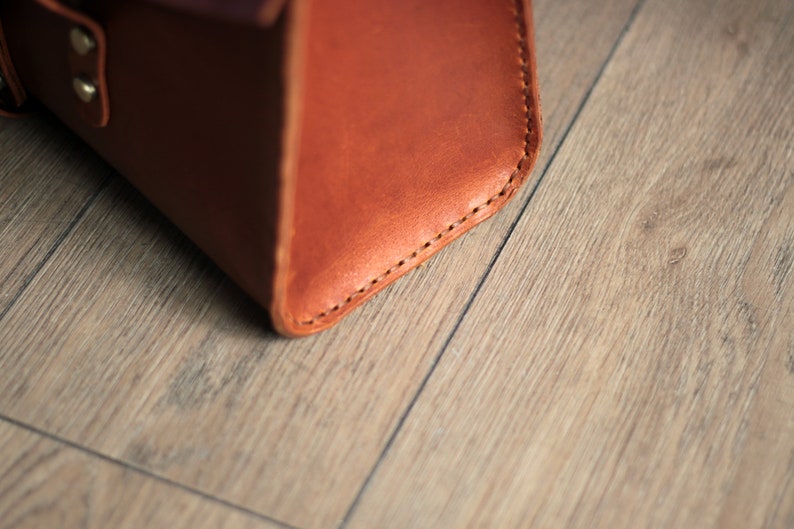 May include: Close-up of the bottom corner of a brown leather bag with stitching detail.