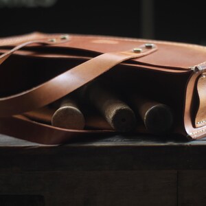 Puede incluir: Una bolsa de herramientas de cuero marr&oacute;n con asa y bandolera. La bolsa es rectangular con esquinas redondeadas y contiene varias herramientas de madera. La bolsa est&aacute; sobre una superficie de madera oscura.