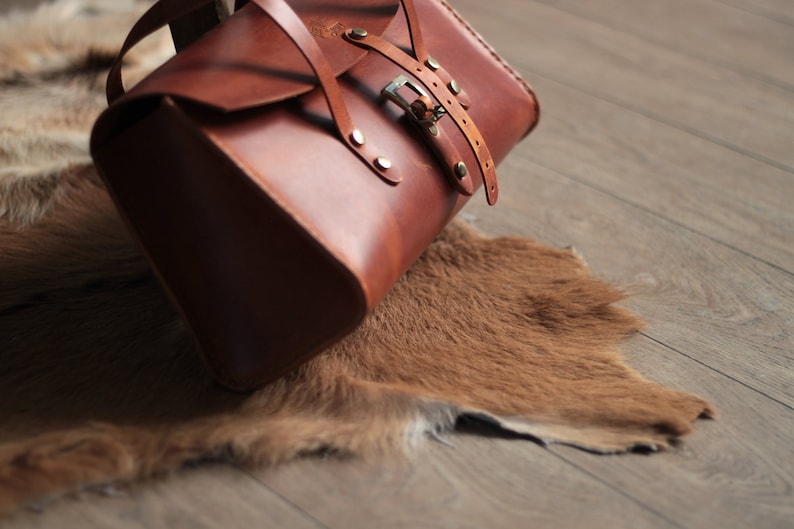 May include: A brown leather satchel bag with a buckle closure and a strap. The bag is sitting on a brown fur rug.