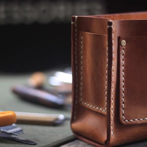 Puede incluir: Primer plano de un organizador de cuero marr&oacute;n con costuras blancas y un peque&ntilde;o bolsillo. El organizador rectangular est&aacute; dise&ntilde;ado para guardar varios objetos. Se ven utensilios en el fondo.