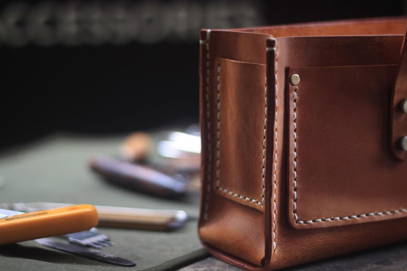 Puede incluir: Primer plano de un organizador de cuero marr&oacute;n con costuras blancas y un peque&ntilde;o bolsillo. El organizador rectangular est&aacute; dise&ntilde;ado para guardar varios objetos. Se ven utensilios en el fondo.