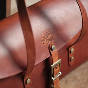 May include: A brown leather satchel bag with a buckle closure and a strap. The bag has a logo embossed on the front that reads "The Mill Story".