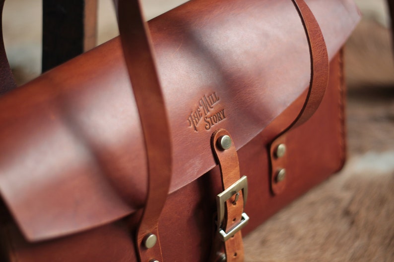 May include: A brown leather satchel bag with a buckle closure and a strap. The bag has a logo embossed on the front that reads "The Mill Story".