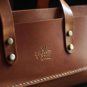 Puede incluir: Primer plano de un bolso tote de cuero marr&oacute;n con dos asas y remaches met&aacute;licos. El bolso tiene el texto en relieve "The Mill Story" y costuras blancas a lo largo del borde inferior. El cuero tiene un acabado liso y pulido.