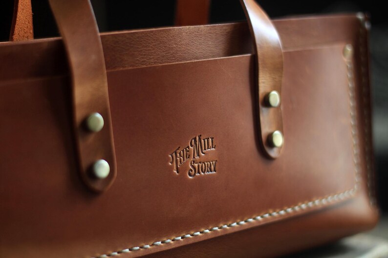 Puede incluir: Primer plano de un bolso tote de cuero marr&oacute;n con dos asas y remaches met&aacute;licos. El bolso tiene el texto en relieve "The Mill Story" y costuras blancas a lo largo del borde inferior. El cuero tiene un acabado liso y pulido.