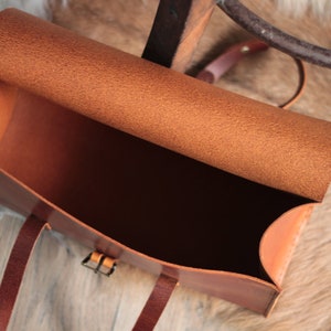 May include: A brown leather satchel bag with a flap top and a buckle closure. The bag is open and the inside is empty.