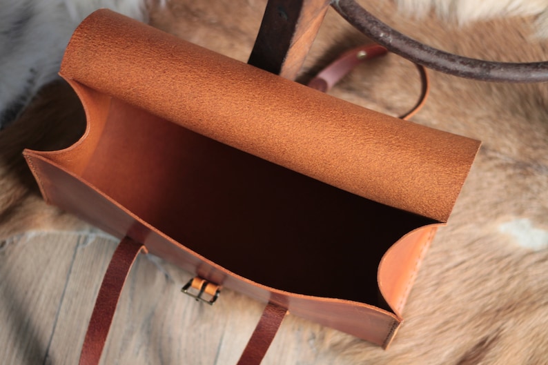 May include: A brown leather satchel bag with a flap top and a buckle closure. The bag is open and the inside is empty.