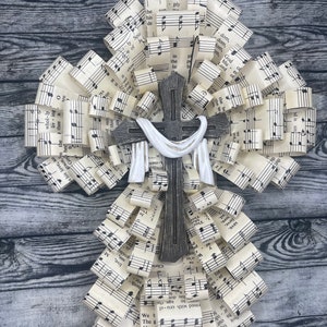 May include: A wooden cross with a white fabric scarf draped over it, surrounded by a wreath of rolled up sheet music. The music sheets have musical notes and lyrics, including the words "God which" and "Lord, which".