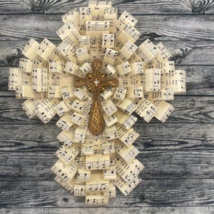 May include: A decorative cross made from folded sheet music, featuring a gold-colored cross in the center. The cross is embellished with a small, clear gem. The background is a weathered, gray wooden surface.