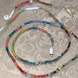 Könnte beinhalten: Ohrhörer mit einem farbenfrohen, dekorativen Kabel. Das Kabel ist mit Fäden in verschiedenen Farben umwickelt, darunter Rot, Orange, Grün, Blau und Lila. Die Ohrhörer und der Ladeanschluss sind weiß.