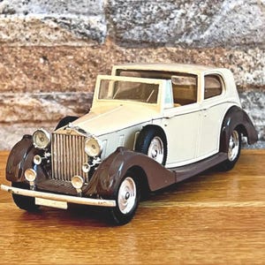 Könnte beinhalten: Ein cremefarbenes und braunes Oldtimer-Modellauto mit detaillierten Scheinwerfern und einem klassischen Kühlergrill. Das Auto steht vor einem ziegelartigen Hintergrund und einer Holzoberfläche.
