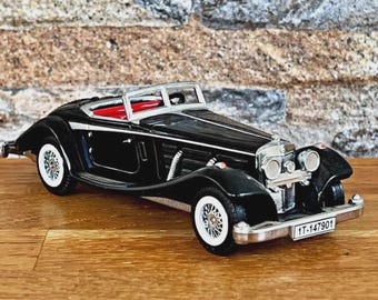 Op de afbeelding: Een zwart vintage cabrioletmodel met whitewall-banden en een rood interieur. De auto heeft zilveren sierlijsten en een kentekenplaat met "1T-147901". Het model staat op een houten oppervlak.