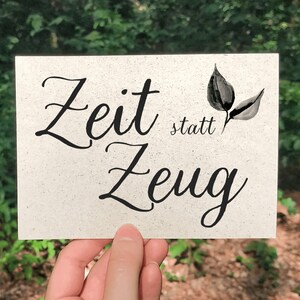 Könnte beinhalten: Eine weiße Karte mit schwarzem Text, der "Zeit statt Zeug" lautet, mit einem kleinen schwarzen Blattdesign. Die Karte wird von einer Hand vor einem verschwommenen grünen Hintergrund gehalten.