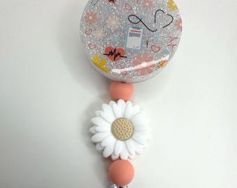 May include: A white daisy charm with a pink and white beaded retractable badge holder. The badge holder has a round, clear, glitter-covered charm with a pink and white floral pattern and medical symbols.