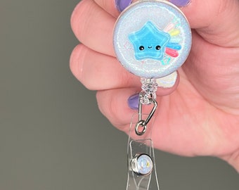 Könnte beinhalten: Ein weißer Glitzer-Badge-Rollenhalter mit einem blauen Stern mit einem Regenbogen und einem niedlichen Gesicht. Der Rollenhalter hat einen durchsichtigen Plastikclip und einen silbernen Drehverschluss.