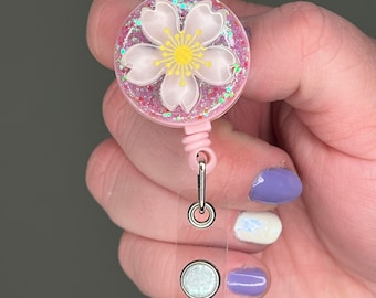 May include: Pink glitter badge reel with a white cherry blossom design. The reel has a clear plastic badge holder with a white glitter circle.
