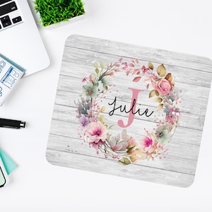 May include: A personalised mouse mat with a floral wreath design on a whitewashed wood background. The wreath features pink, green and purple flowers. The initial "J" is pink and the name "Julie" is written in black script.