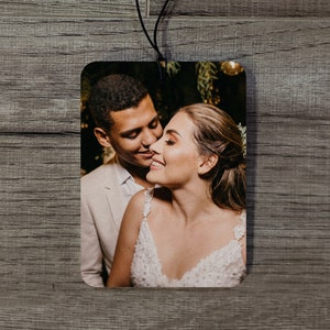 May include: Rectangular air freshener featuring a color photograph of a smiling couple. The man wears a light-colored suit jacket, and the woman is in a white dress. A black string is attached for hanging.