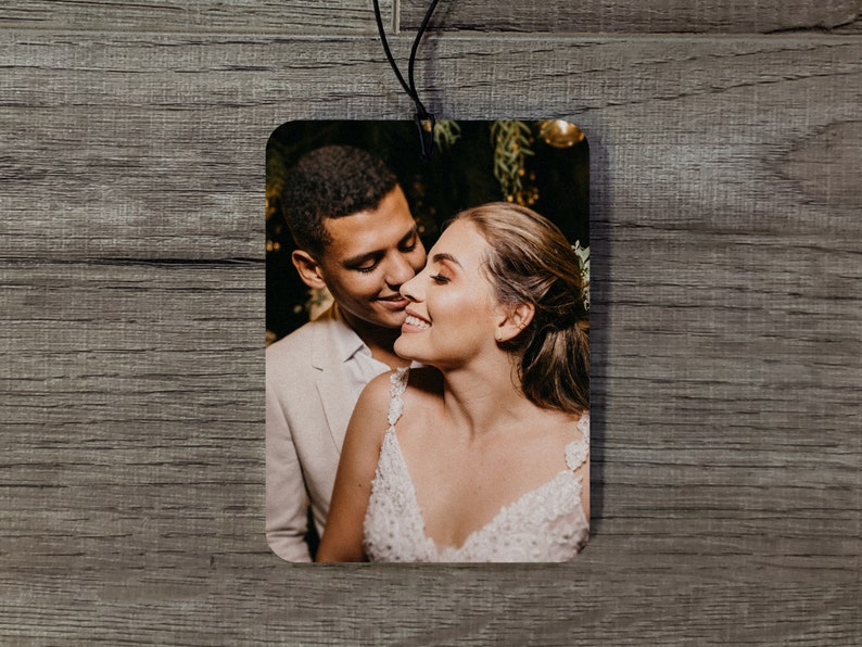 May include: Rectangular air freshener featuring a color photograph of a smiling couple. The man wears a light-colored suit jacket, and the woman is in a white dress. A black string is attached for hanging.