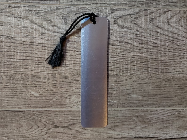 Puede incluir: Un marcap&aacute;ginas de metal plateado con un borla negra. El marcap&aacute;ginas est&aacute; sobre una superficie de madera.