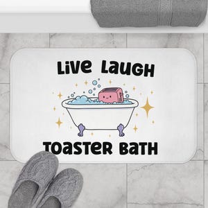 May include: White rectangular bath mat featuring the words "Live Laugh" above a cartoon bathtub with a pink toaster inside. The words "Toaster Bath" are below the bathtub. Grey knitted slippers are at the bottom.
