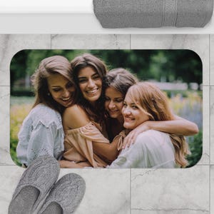 May include: Rectangular bath mat featuring a photograph of four smiling women embracing. The mat has rounded corners and is placed on a tiled floor. Gray slippers are in the foreground, and a gray towel is at the top.