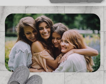 May include: Rectangular bath mat featuring a photograph of four smiling women embracing. The mat has rounded corners and is placed on a tiled floor. Gray slippers are in the foreground, and a gray towel is at the top.