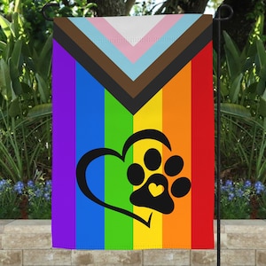 May include: A rainbow pride flag with a brown, light blue, and pink triangle in the top left corner. A black paw print with a yellow heart inside is in the center of the flag.