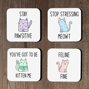 May include: Four white square coasters with cat illustrations and puns. The coasters feature the text "Stay Pawsitive", "Stop Stressing Meowt", "You've Got To Be Kitten Me", and "Feline Fine".