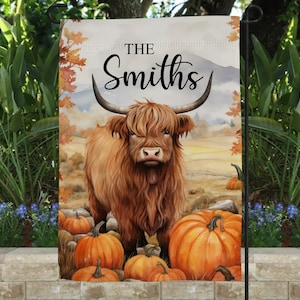 May include: A rectangular garden flag featuring a brown Highland cow standing in a field with orange pumpkins. The text "THE Smiths" is printed in black script and serif font at the top. The background shows a landscape with autumn foliage.