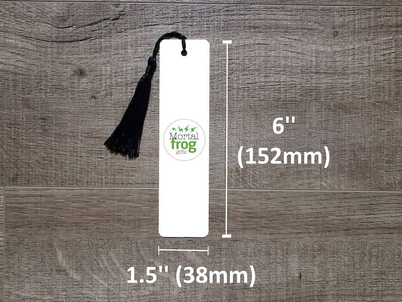 Puede incluir: Marcap&aacute;ginas blanco con un borla negra y el texto "Mortal Frog Gifts" en un c&iacute;rculo verde. El marcap&aacute;ginas mide 15,2 cm de alto y 3,8 cm de ancho.