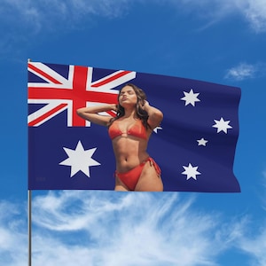 May include: A woman in a red bikini stands before an Australian flag against a blue sky. The flag shows the Union Jack and the Southern Cross constellation. The flag is waving in the wind.