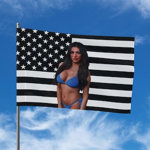 Könnte beinhalten: Eine Flagge mit einem schwarz-weißen amerikanischen Flaggenmuster, mit einer Frau in einem blauen Bikini, die auf die Flagge gelegt wurde. Die Flagge befindet sich auf einer Metallstange vor einem blauen Himmel mit Wolken.