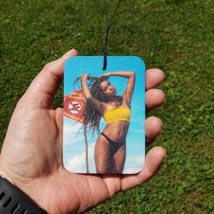 May include: Rectangular air freshener featuring a woman in a yellow top and black bottoms, with a beach scene background. The image includes a red sign with text. The air freshener is held against a green grass background.