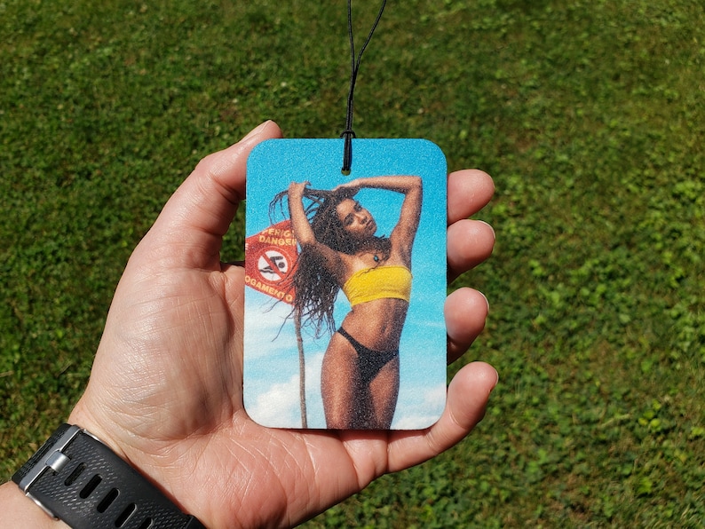 May include: Rectangular air freshener featuring a woman in a yellow top and black bottoms, with a beach scene background. The image includes a red sign with text. The air freshener is held against a green grass background.