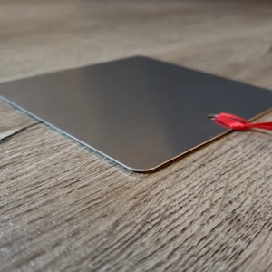May include: A silver metal square tag with a red ribbon attached. The tag is blank and has a hole punched in the top corner.