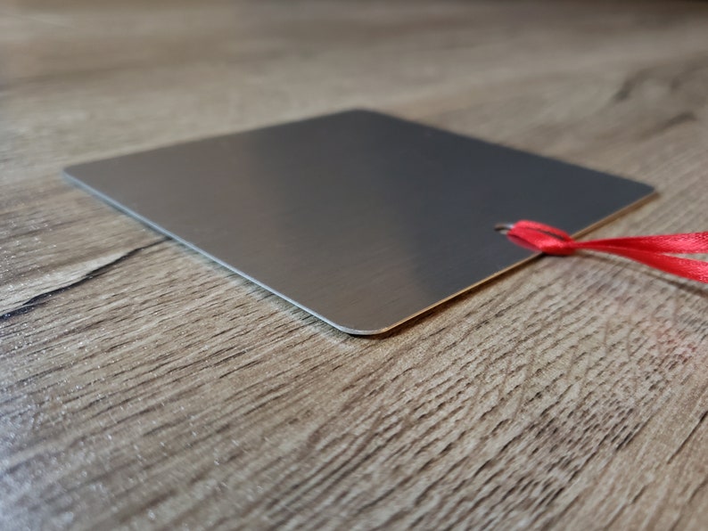 May include: A silver metal square tag with a red ribbon attached. The tag is blank and has a hole punched in the top corner.