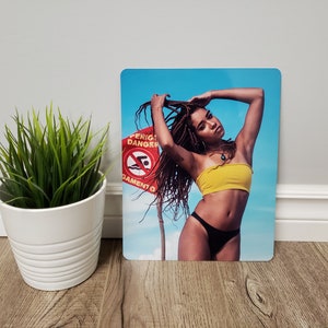 May include: A woman with long braids poses in a yellow bandeau top and black bikini bottoms. She is standing in front of a blue sky with a red and white sign that says "Perigo Danger" and "Nata&ccedil;&atilde;o Proibida".