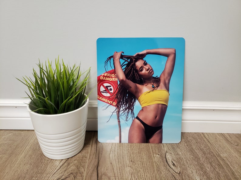 May include: A woman with long braids poses in a yellow bandeau top and black bikini bottoms. She is standing in front of a blue sky with a red and white sign that says "Perigo Danger" and "Nata&ccedil;&atilde;o Proibida".