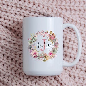 May include: White ceramic mug with a floral wreath design, the name "Julie", and the letter "J". The wreath features pink, green, and peach flowers. The mug has a large handle and is set against a pink knitted background.