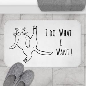 May include: White rectangular bath mat with a black cat illustration and the text "I DO WHAT I WANT!". The cat is drawn in a playful pose. Gray knitted slippers are at the bottom of the mat.