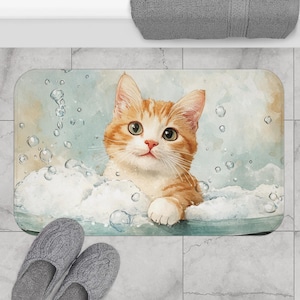 May include: A rectangular bath mat with a watercolour illustration of a ginger and white kitten in a bubble bath. The mat has a light blue and white colour scheme with bubbles and a smiling cat.