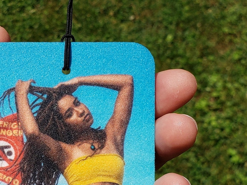 May include: A rectangular air freshener with a woman in a yellow tube top and turquoise necklace against a blue background. The woman is holding her hair. A black string is attached to the top.