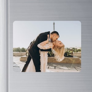 May include: A couple in love, the woman in a white wedding dress, is being lifted up by the man in a black suit. They are kissing in front of the Eiffel Tower in Paris, France.